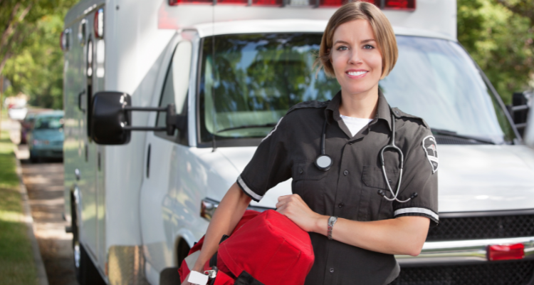 A smiling paramedic in uniform stands in front of an ambulance, holding a red medical bag, with a stethoscope around their neck and trees visible in the background