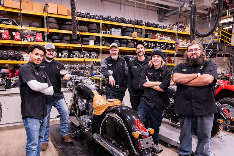 Students and instructors are gathered around a motorcycle in a small engine repair lab.