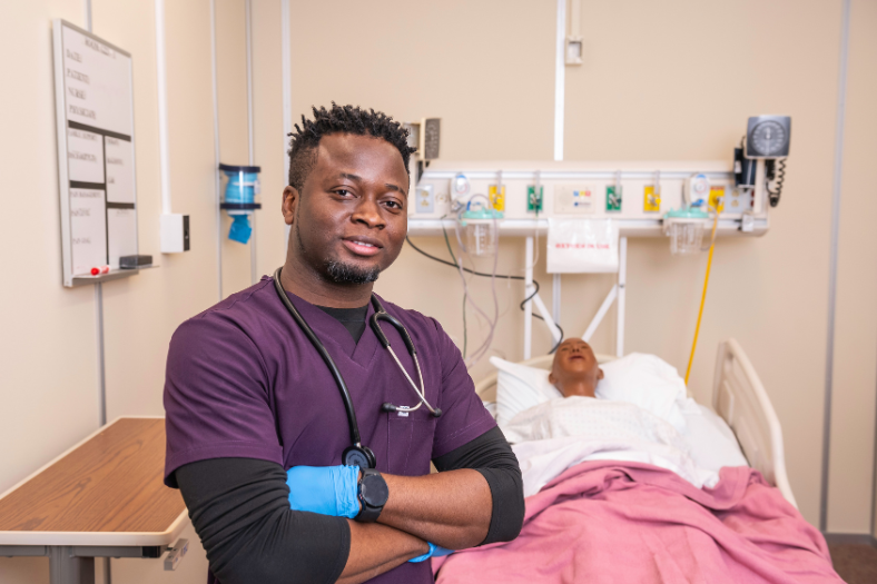 An student stands wearing purple scrubs and blue gloves with their arms folded. There is a patient bed in the background.