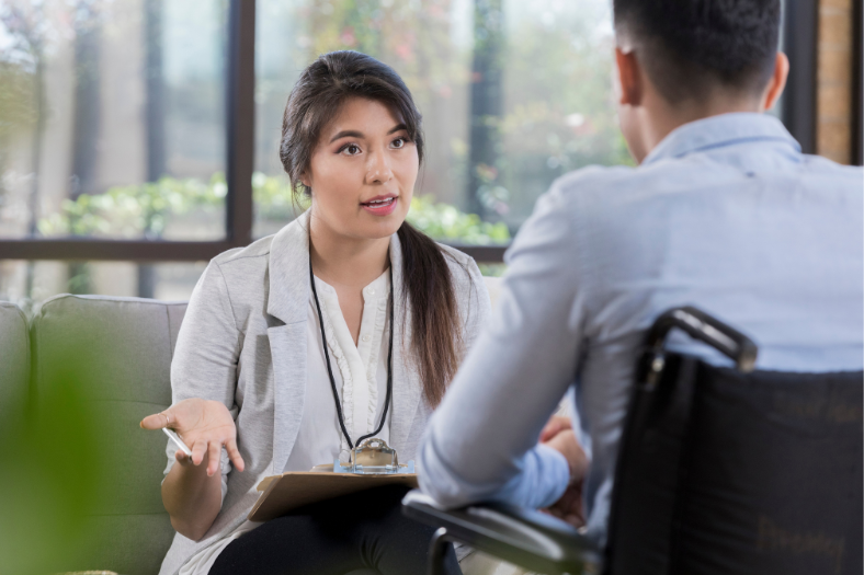 A young professional woman holding a clipboard engages in a conversation with a man using a wheelchair, seated across from her in a brightly lit space.